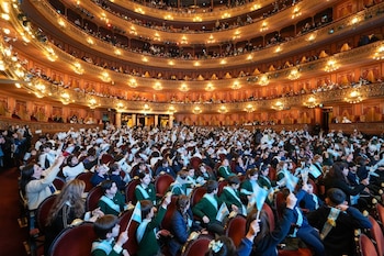 Numerosos niños y adultos en plateas y palcos de un teatro. Muchos niños agitan pequeñas banderas argentinas