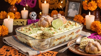 Recipiente de vidrio con postre de limón y pan de muerto, acompañado de una porción servida, velas encendidas y flores de cempasúchil.
