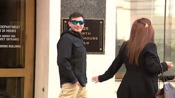 Hombre sonriente con gafas de sol, chaqueta oscura y pantalones claros, camina junto a una mujer de espaldas frente a un edificio