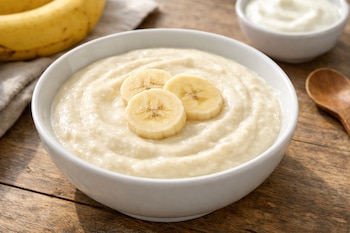 Un plato hondo blanco lleno de crema suave de plátano y yogur, adornado con tres rodajas de plátano. La mesa de madera recibe luz natural.
