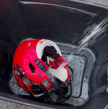 Un casco de fútbol americano rojo con detalles blancos se lava con un chorro de agua en un recipiente negro