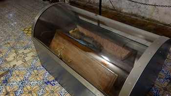 Vitrina de cristal con borde metálico contiene un ataúd de madera con un crucifijo dorado. En el interior se observa una momia. El suelo es de azulejos