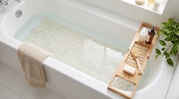 Bañera blanca con agua, alfombrilla de baño circular, toalla beige, bandeja de bambú con jabón y cepillo, botellas de producto y planta.
