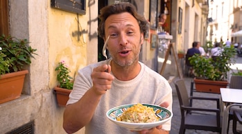Un hombre de mediana edad sostiene un plato de comida y un tenedor frente a una calle con edificios antiguos, macetas y mesas.