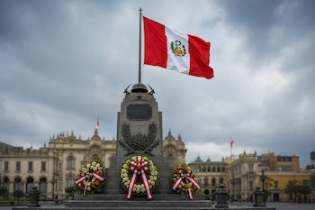 Monumento de piedra con la bandera de Perú, una placa y arreglos florales con cintas rojas y blancas ante edificios históricos.
