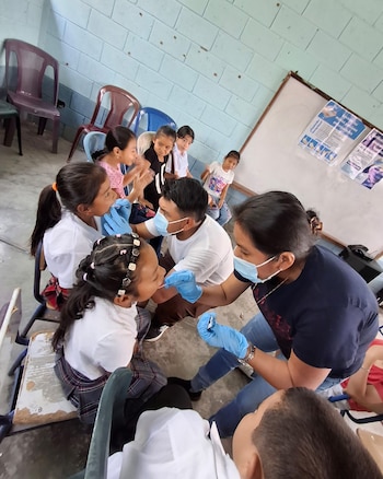 Profesionales de la salud con mascarillas y guantes azules examinan la boca de varios estudiantes en un aula de escuela, con niños observando