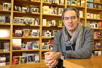 Hombre de mediana edad con gafas y bufanda gris, apoyado en una mesa de madera. Detrás, estanterías de madera con libros y marcos de fotos