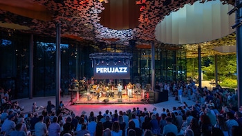 Banda musical en escenario con pantalla que muestra "PERUJAZZ". Público sentado, paredes de vidrio y techo de diseño. Fondo de árboles nocturnos
