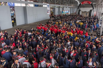 Vista aérea de un gran grupo de estudiantes en uniformes escolares, principalmente azul marino y rojo, en un concurrido pabellón de exposiciones