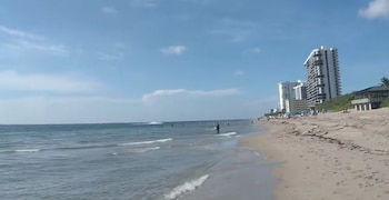 Una playa de arena con personas, oleaje en el mar y edificios de varios pisos al fondo bajo un cielo azul