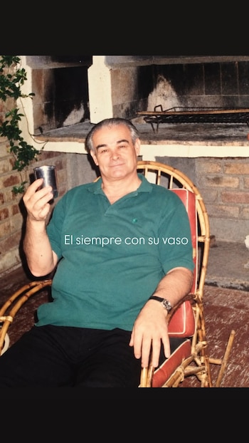 Un hombre mayor con polo verde sostiene un vaso de metal sentado en una silla de mimbre frente a una parrilla