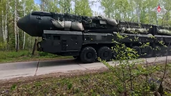 Un vehículo militar pesado de color verde oscuro, con un gran misil cubierto por camuflaje y sacos de arena, circula por una carretera rodeada de árboles en un bosque