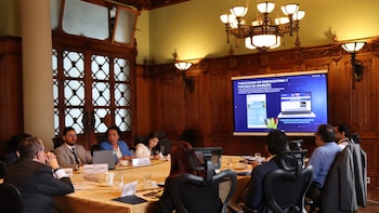 Vista de una sala de reuniones con varias personas sentadas alrededor de una mesa grande, frente a una pantalla que muestra información digital