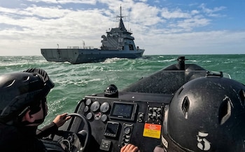 Vista desde una lancha rápida con un marinero al volante, observando un gran buque de guerra gris en movimiento en un mar picado bajo un cielo parcialmente nublado