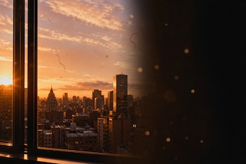 Vista de una ciudad al atardecer desde una ventana con una sombra lateral y formas traslúcidas flotantes en el encuadre.