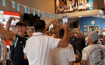 Un grupo de personas en un local con banderas de Argentina y un mural de un equipo de fútbol. Se observan televisores, un reloj de pared y un teléfono móvil