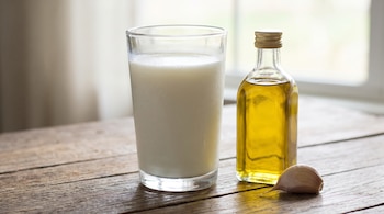 Un vaso de leche entera fría, una botella de aceite de oliva y un diente de ajo se ubican sobre una superficie de madera, con un fondo claro.