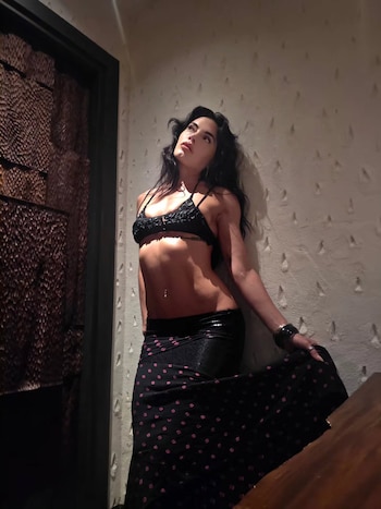 Emilia Attias posa de pie vestida con un top negro y una falda larga estampada junto a una pared clara y una puerta de madera