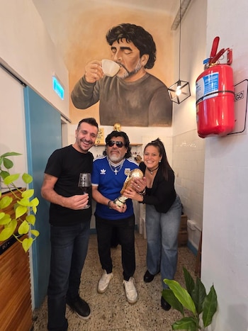 Un hombre con una copa, un imitador de Diego Maradona y una mujer sostienen la réplica de un trofeo bajo el mural de un hombre tomando café
