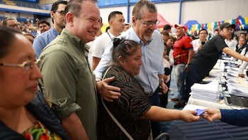 Un hombre habla frente a un podio junto a la bandera de Guatemala ante una pantalla con el anuncio de la tarjeta 75.000 del Programa Bolsa Social