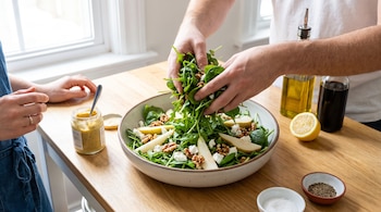 Manos colocando rúcula en un cuenco con rodajas de pera, queso y nueces junto a aceite, vinagre, limón y frasco de mostaza sobre madera.
