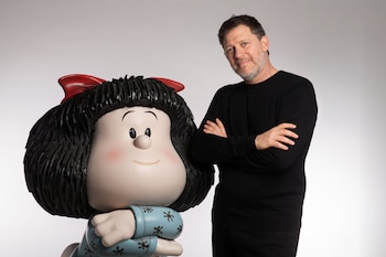 Un hombre de barba y suéter negro posa de pie junto a una figura de Mafalda con pelo negro, moño rojo y blusa azul