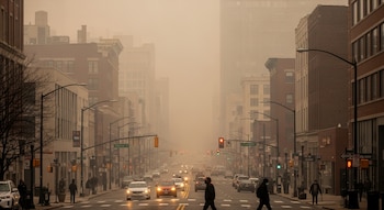 Vista de una calle urbana con edificios altos y tráfico moderado, envuelta en una densa bruma de smog gris y naranja. Algunas personas cruzan la calle usando mascarillas.