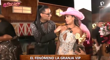 Shirley Arica con vestido plateado brillante, sombrero vaquero rosa y un trofeo de gallo dorado, conversando con una persona con camisa de lentejuelas en un estudio de televisión