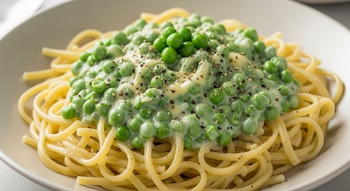 Un plato hondo contiene fideos cubiertos con una salsa cremosa de arvejas verdes, queso rallado y pimienta negra molida. El fondo es neutro.