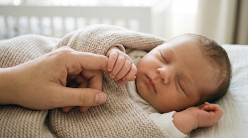 Un bebé recién nacido duerme tranquilamente, envuelto en una manta beige de punto, con su pequeña mano agarrando el dedo de una persona adulta.