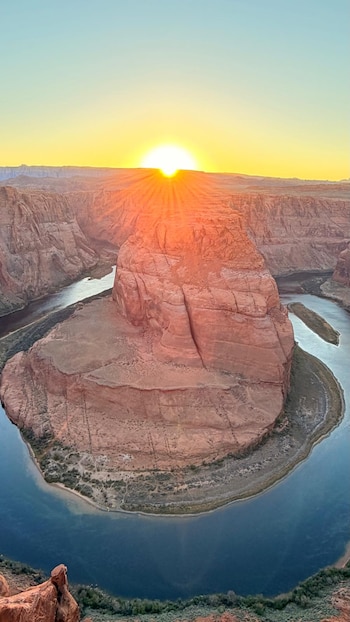 Formación rocosa rodeada por un cauce de agua en curva con el sol en el horizonte