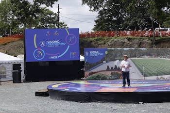 Un hombre sobre una tarima circular junto a una pantalla digital y un cartel informativo del proyecto Ciudad Deportiva Los Amates