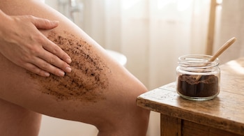 Primer plano de una persona aplicando un exfoliante de café oscuro en su muslo. A la derecha, un frasco de vidrio con café molido y una cuchara de madera sobre una mesa de madera.
