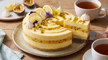 Torta de mousse de limón y maracuyá en plato, con rodajas de limón, merengues, flores y fruta de la pasión. Una porción es servida. Hay dos tazas de té.