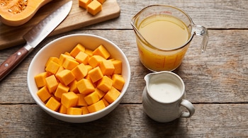 Media calabaza, cuchillo, tabla de madera, un cuenco con cubos de zapallo, una jarra medidora con caldo de verduras y una jarra pequeña con crema de leche sobre madera.