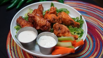 Primer plano de un plato blanco con alitas de pollo cubiertas en salsa, lechuga, dos recipientes pequeños con aderezo blanco, y bastones de apio y zanahoria