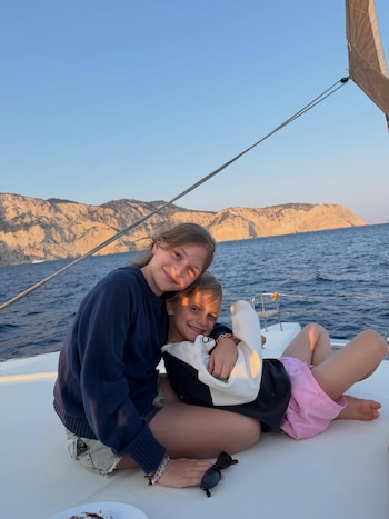 Primer plano de dos niños sonrientes en el lateral de un barco. Uno lleva jersey azul y shorts de jean, el otro sudadera y shorts rosas. De fondo, mar y montaña