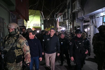 Grupo de hombres, incluyendo policías con uniforme y equipo táctico, caminan por una calle estrecha de noche; algunos edificios visibles con poca luz