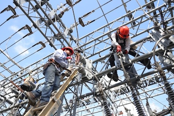 Dos electricistas con cascos rojos y arneses trabajan en una estructura metálica con cables e aisladores eléctricos bajo un cielo azul claro, uno de ellos sobre una escalera