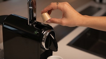 Mano inserta cápsula de café beige en máquina de café negra con detalles cromados y botón verde, sobre encimera de cocina.