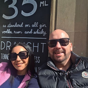 Hombre calvo con barba y gafas de sol, mujer con pelo oscuro y gafas de sol. Detrás, cartel de pizarra negro con texto sobre bebidas alcohólicas