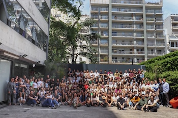 Un numeroso grupo de personas posa al aire libre en un patio de cemento. Se ven hombres y mujeres de diversas edades frente a un edificio, follaje y otros edificios altos al fondo