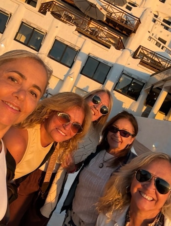 Cinco mujeres adultas sonríen directamente a cámara, cuatro de ellas con gafas de sol, frente a un edificio blanco de múltiples balcones y ventanas