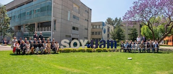 Vista exterior del campus de la UAM con un gran grupo de personas sentadas en el césped, flanqueando un letrero blanco y azul que dice "SOY UAM". Se ven edificios modernos y árboles al fondo