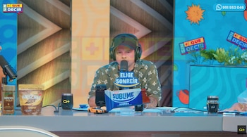 Un hombre con gorra y auriculares habla frente a un micrófono en un estudio de televisión con un empaque de chocolate sobre la mesa