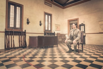 Hombre con traje claro y bigote sentado en una silla de madera en un cuarto con piso de ajedrezado. Hay rifles en un estante junto a una ventana