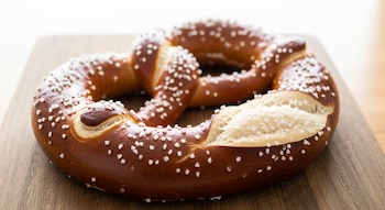 Un pretzel grande y dorado con corteza brillante y sal gruesa visible, reposa sobre una tabla rústica de madera con un fondo claro y difuso.
