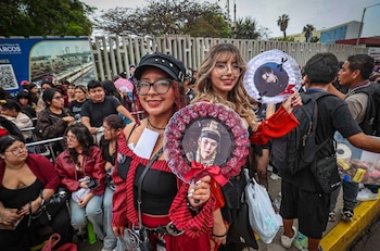 Dos mujeres sostienen abanicos con retratos impresos frente a una multitud de personas reunidas en la calle junto a una reja