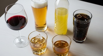 Copa de vino, vaso de cerveza, botella de bebida energética, vaso de gaseosa oscura con hielo, vaso de whisky con hielo y vaso de licor ámbar sobre una mesa.