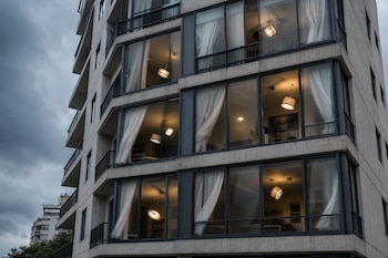 Fachada de un edificio de concreto con grandes ventanas, cortinas blancas, lámparas de techo encendidas y cielo nublado.